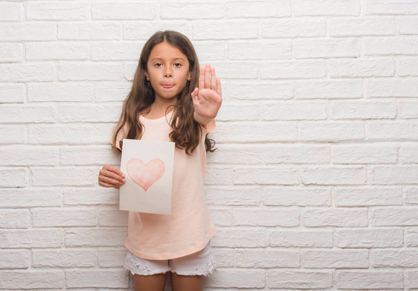 Jovem criança hispânica sobre parede de tijolo branco dando cartão de dia mãe com a mão aberta fazendo sinal de parada com expressão séria e confiante, gesto de defesa
 - Foto, Imagem