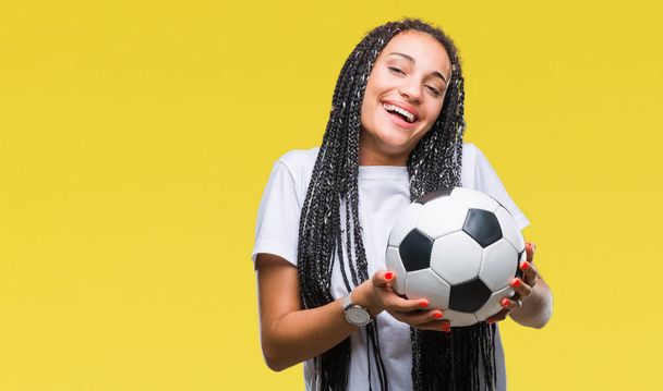 Joven chica afroamericana trenzada pelo sosteniendo pelota de fútbol sobre un fondo aislado con una cara feliz de pie y sonriendo con una sonrisa confiada mostrando los dientes
 - Foto, imagen