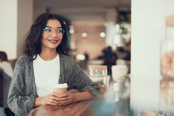 Cute young lady holding cup of coffee and smiling while sitting alone - Foto, afbeelding