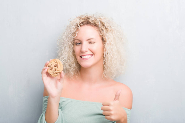 Молода блондинка над гранжевою сірою стіною їсть шоколадний чіп з кулінарним задоволенням з великою посмішкою роблячи знак ОК, великий палець з пальцями, відмінний знак
 - Фото, зображення