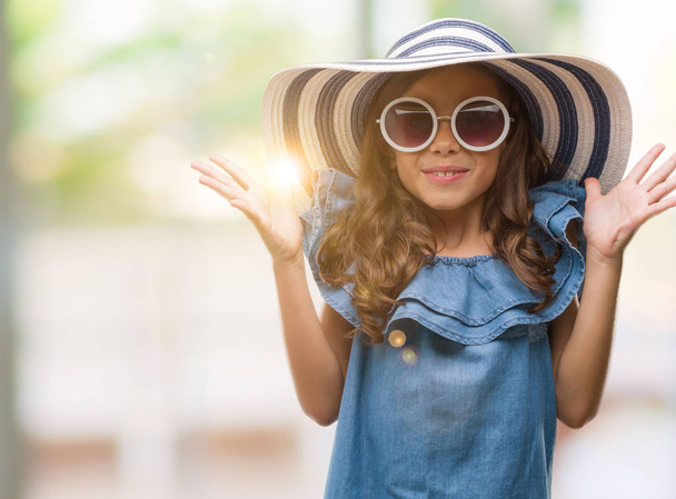 Brunette hispanic girl wearing sunglasses and summer hat very happy and excited, winner expression celebrating victory screaming with big smile and raised hands - Photo, image
