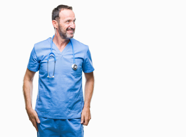 Középkorú Deres vezető orvos férfi orvosi egységes visel át elszigetelt háttér keres el az oldalon mosollyal arcán, természetes kifejezése. Nevetve magabiztos. - Fotó, kép
