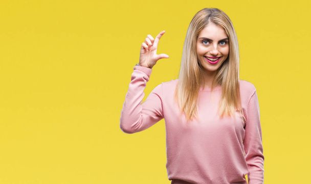 Young beautiful blonde woman wearing pink winter sweater over isolated background smiling and confident gesturing with hand doing size sign with fingers while looking and the camera. Measure concept. - Foto, Imagen