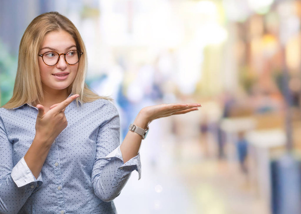 Jonge Kaukasische zakenvrouw bril over geïsoleerde achtergrond verbaasd en lachend naar de camera tijdens het presenteren met hand en met de vinger te wijzen. - Foto, afbeelding