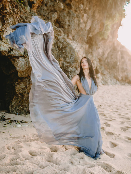 Young woman posing in stylish blue dress in beach at sunset or sunrise colors - Photo, Image