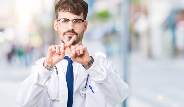 Nuori lääkäri mies yllään sairaalan takki yli eristetty tausta hylkääminen ilme ristikkäin sormet tekee negatiivinen merkki
 - Valokuva, kuva