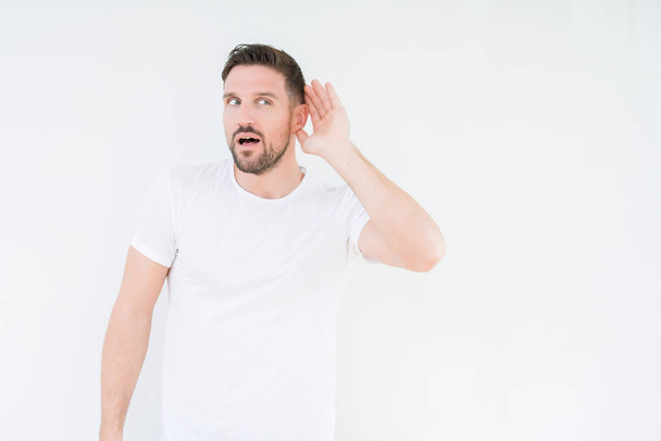Молодой красивый мужчина в обычной белой футболке на изолированном фоне улыбается рукой над ухом, слушая слухи или сплетни. Концепция глухоты
. - Фото, изображение