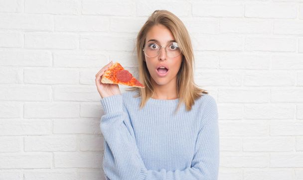 驚き顔、恐れ、恐怖表現と興奮とショックで怖いピザのスライスを食べる白いレンガの壁に美しい若い女性 - 写真・画像