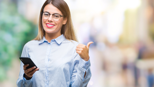 Young beautiful business woman texting message using smartphone over isolated background pointing and showing with thumb up to the side with happy face smiling - Foto, afbeelding