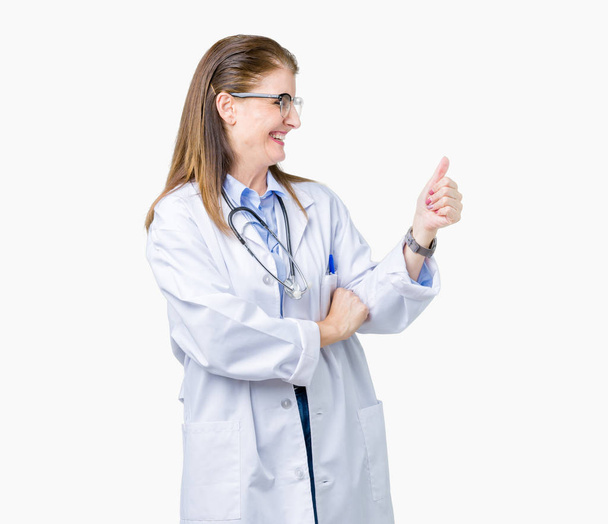 Średnim wieku starsze lekarza kobieta uśmiechając się robi sobie płaszcz medyczny na białym tle patrząc dumny, kciuk w górę gest w bok - Zdjęcie, obraz