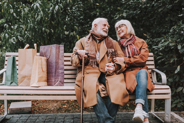 Happy elderly couple sitting on bench with shopping bags - 写真・画像