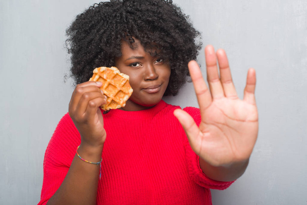 Jeune femme afro-américaine sur un mur gris grunge mangeant une gaufre belgique à la main ouverte faisant signe d'arrêt avec expression sérieuse et confiante, geste de défense
 - Photo, image