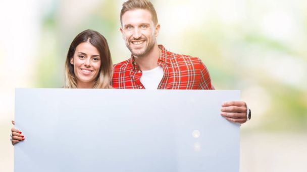Genç çift birlikte holding boş afiş üzerinde duran ve diş gösterilen kendine güvenen bir gülümsemeyle gülümseyerek mutlu bir yüz ile izole arka plan - Fotoğraf, Görsel
