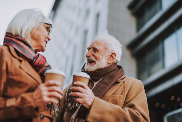 Side view portrait of stylish old gentleman in coat chatting with wife. They holding cups of coffee while looking at each other and smiling - Foto, immagini