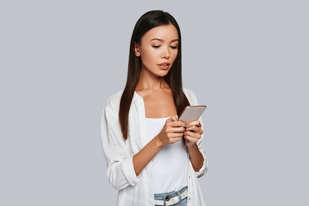 Beautiful young Asian woman using her smart phone while standing against grey background - Φωτογραφία, εικόνα