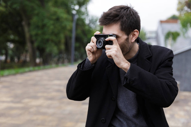 Photo of brunette man 20s wearing black coat photographing on retro camera while walking outdoor - Φωτογραφία, εικόνα