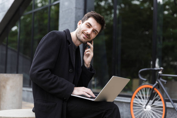 Image of handsome young business man sitting outdoors using laptop computer talking by phone. - Foto, Bild