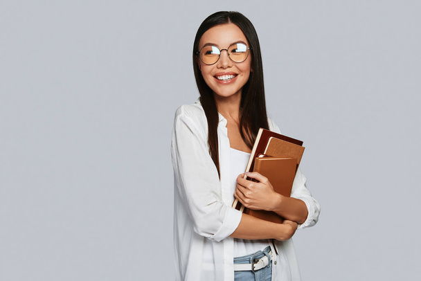 Beautiful young Asian woman carrying books and smiling while standing against grey background - 写真・画像