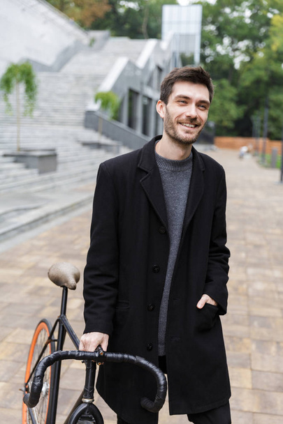 Happy young man dressed in coat walking with a bicycle on a street - 写真・画像