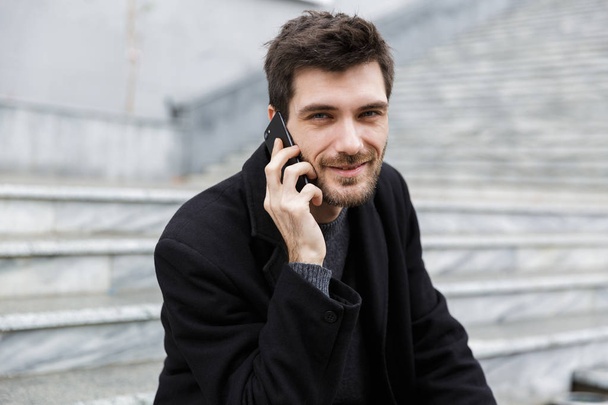Smiling man dressed in coat sitting at the city street, talking on mobile phone - Φωτογραφία, εικόνα