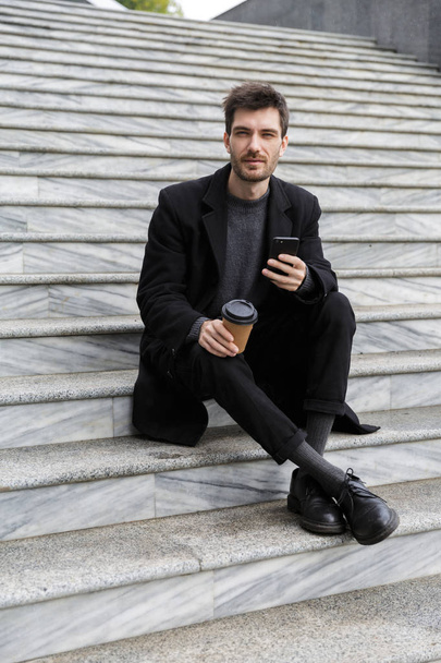 Image of handsome young business man sitting outdoors using mobile phone drinking coffee. - Zdjęcie, obraz