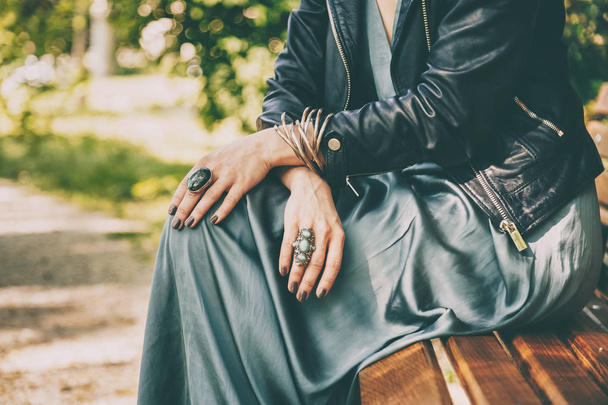fashion detail, close-up of a woman wearing silk dress, leather jacket and rings and bracelets as accessories, while sitting on a bench in a park - Photo, Image