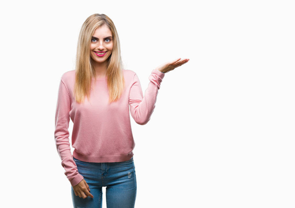 Jovem mulher loira bonita vestindo suéter de inverno rosa sobre fundo isolado sorrindo alegre apresentando e apontando com palma da mão olhando para a câmera
. - Foto, Imagem