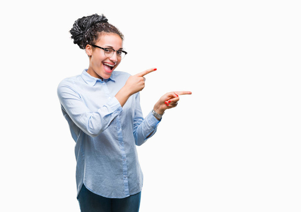 junge geflochtene Haare afrikanisch-amerikanische Geschäftsfrau mit Brille über isoliertem Hintergrund lächelt und blickt in die Kamera, die mit zwei Händen und Fingern zur Seite zeigt. - Foto, Bild