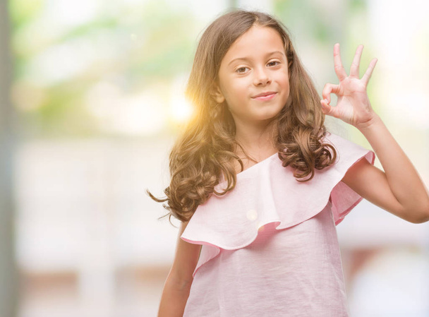 Брюнетка латиноамериканка в розовом платье улыбается позитивно делает хорошо знак с рукой и пальцами. Успешное выражение
. - Фото, изображение