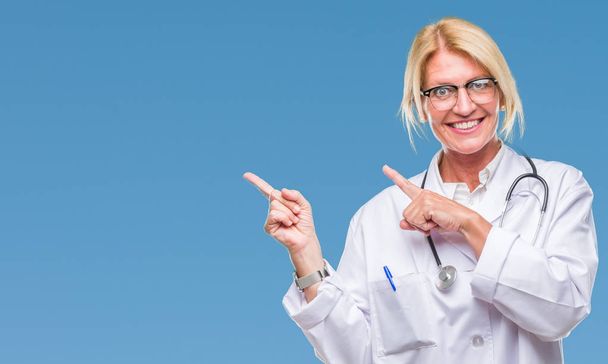 Blonde Ärztin mittleren Alters über isoliertem Hintergrund, lächelnd und mit zwei Händen und Fingern zur Seite in die Kamera blickend. - Foto, Bild
