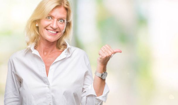 Middle age blonde business woman over isolated background smiling with happy face looking and pointing to the side with thumb up. - Photo, Image