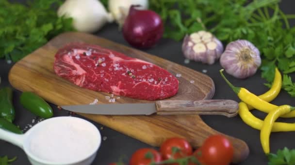 one raw piece of tasty steak with salt, pepper on a wooden board and a set of vegetables on a black table - Materiaali, video