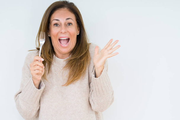 Middle age woman holding silver fork metal over isolated background very happy and excited, winner expression celebrating victory screaming with big smile and raised hands - Valokuva, kuva
