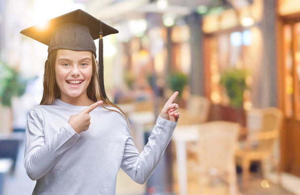 Young beautiful girl wearing graduate cap over isolated background smiling and looking at the camera pointing with two hands and fingers to the side. - Photo, Image