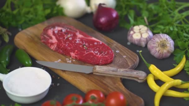 one raw piece of tasty steak with salt, pepper on a wooden board and a set of vegetables on a black table - Záběry, video