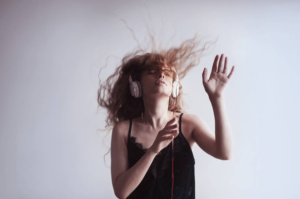 όμορφη νεαρή γυναίκα ακούτε μουσική με ακουστικά μπροστά από το λευκό τοίχο και χορό - Φωτογραφία, εικόνα
