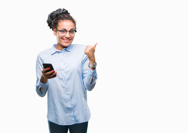 Jonge gevlochten haar Afrikaanse Amerikaanse bedrijfsleven meisje met smartphone over geïsoleerde achtergrond aan te wijzen en te tonen met de duim aan de kant met een blij gezicht lachend - Foto, afbeelding