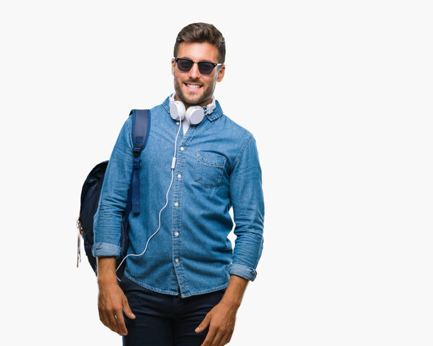 Молодий красивий турист носить навушники та рюкзак над ізольованим тлом, дивлячись на бік з посмішкою на обличчі, природним виразом. Сміється впевнено
. - Фото, зображення