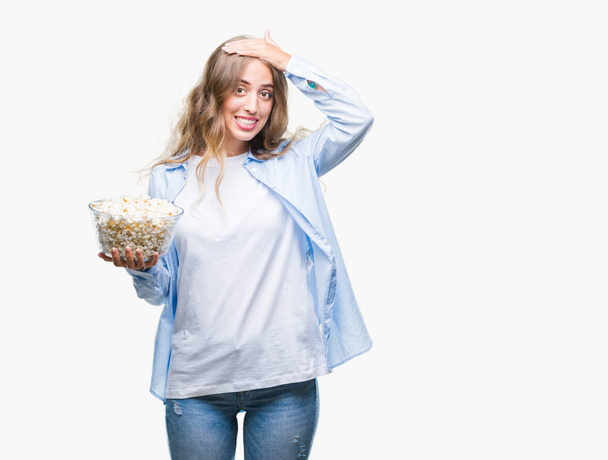 Beautiful young blonde woman eating popcorn over isolated background stressed with hand on head, shocked with shame and surprise face, angry and frustrated. Fear and upset for mistake. - 写真・画像