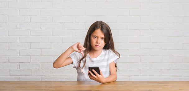 Fiatal spanyol gyerek ül a asztal használ smartphone, haragos arc, negatív előjellel mutatja ellenszenv a hüvelykujját lefelé, elutasítás koncepció - Fotó, kép
