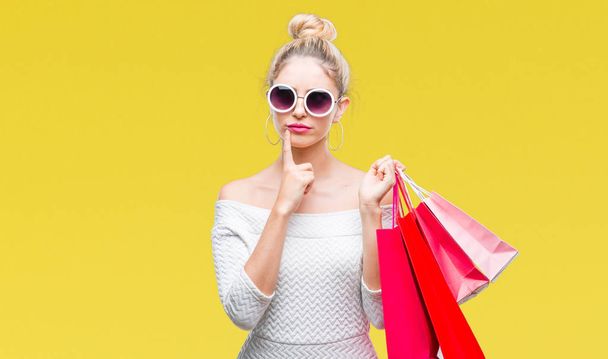 junge schöne blonde Frau mit Einkaufstaschen über isoliertem Hintergrund ernstes Gesicht über Frage nachdenkend, sehr verwirrte Idee - Foto, Bild