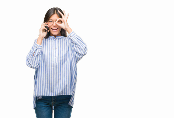 Młoda kobieta azjatyckich, mówiąc na telefon na białym tle z szczęśliwy twarz uśmiechający się robi ok znak ręką na oko patrząc przez palce - Zdjęcie, obraz