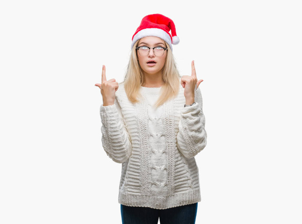 若い白人女性隔離された背景にクリスマスの帽子をかぶってびっくり驚いて見上げると指で指していると腕を上げた. - 写真・画像