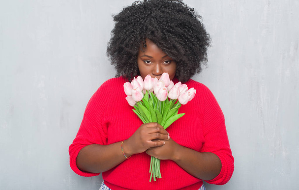 Nuori afrikkalainen amerikkalainen nainen harmaan grunge seinä tilalla vaaleanpunaisia kukkia kimppu luottavainen ilme fiksu kasvot ajattelu vakava
 - Valokuva, kuva