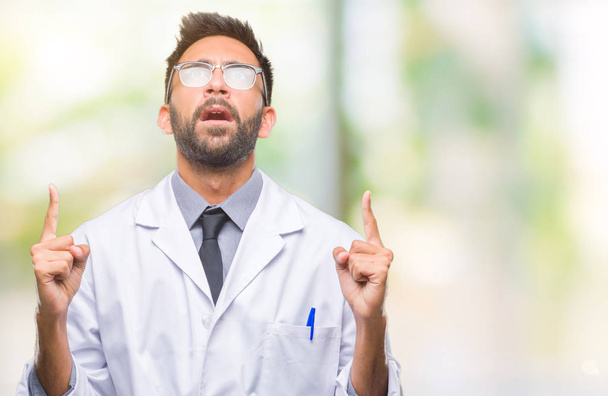 Adult hispanic scientist or doctor man wearing white coat over isolated background amazed and surprised looking up and pointing with fingers and raised arms. - 写真・画像