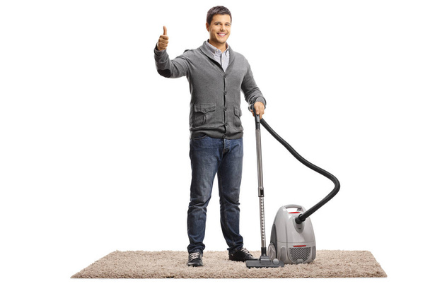 Full length portrait of a young man with a vacuum cleaner on a carpet giving thumbs up isolated on white background - Foto, afbeelding