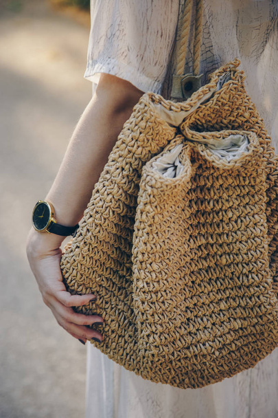 Nahaufnahme einer stilvollen Frau im Sommerkleid, die eine Strohhandtasche in der Hand hält, während sie eine stilvolle Armbanduhr trägt - Foto, Bild
