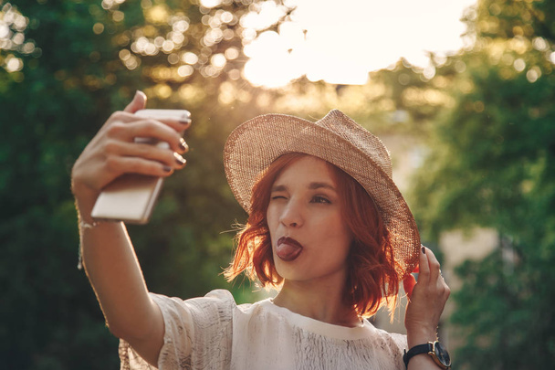Piękne i stylowe czerwone włosy dziewczyna biorąc selfie z jej smartphone na zewnątrz - Zdjęcie, obraz
