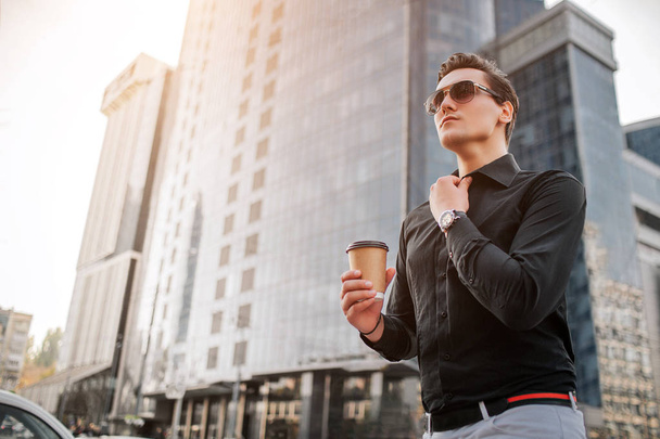 Serious young mn holds hand on collar of shirt and look forward. He has cup of drink. Guy stands in front of skyscrapers. - Foto, Imagem
