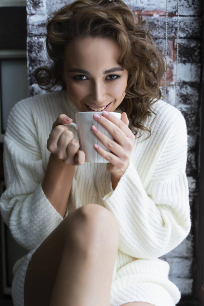 A beautiful girl wearing a white sweater and warm socks sits on the windowsill at the Christmas tree, at a window, behind which is a snow-covered street and drinks a cup of hot drink. Copy space. - Foto, Bild